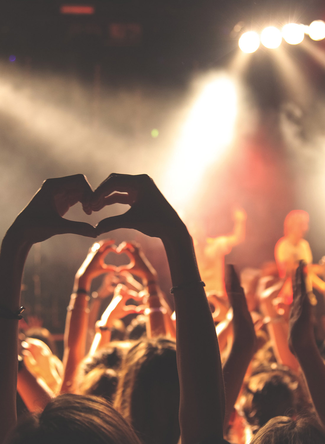 A crowd holds up their hands in the shape of hearts at a live musical performance.